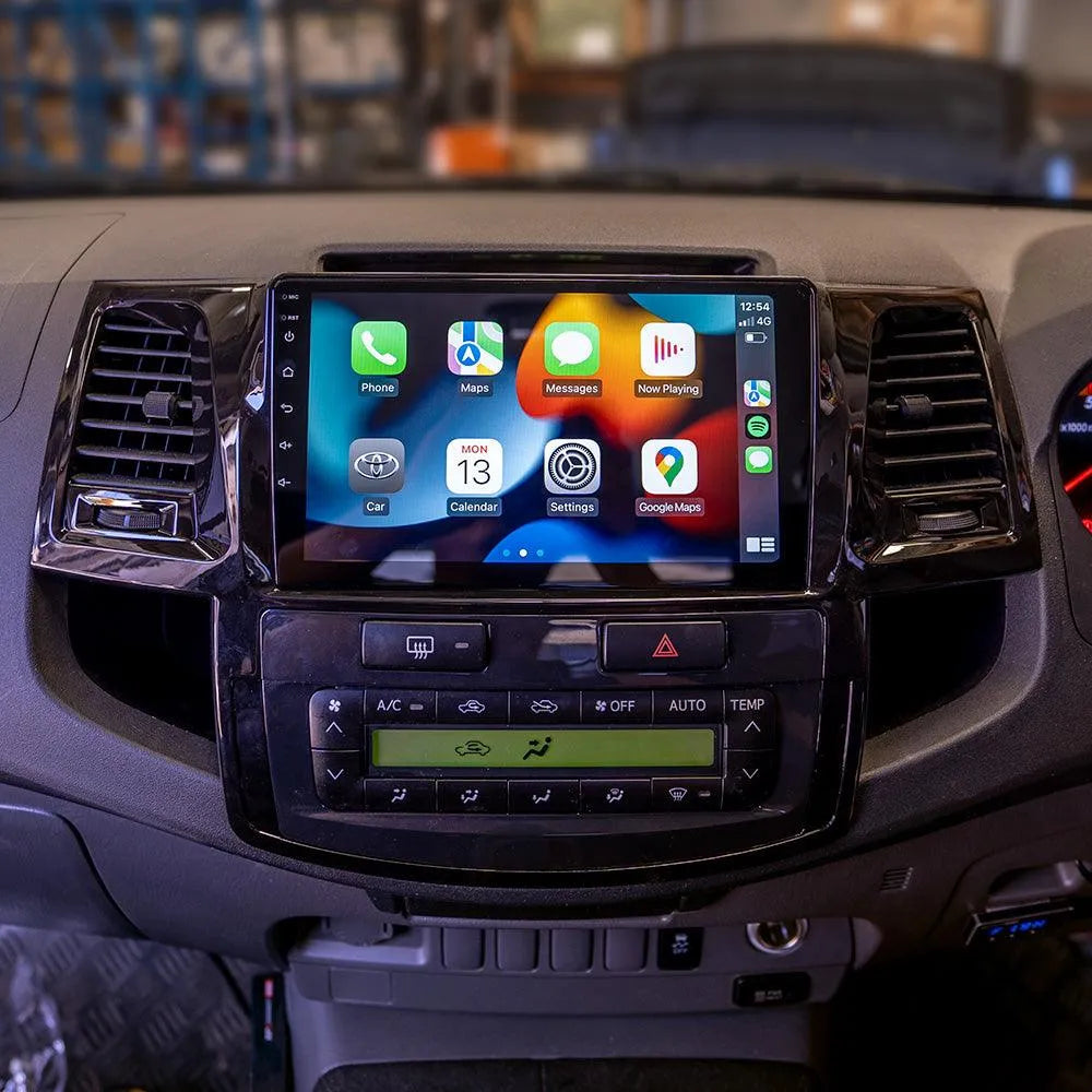 Dashboard of the Toyota Hilux 2005-2015 featuring a multimedia system with Apple CarPlay. The screen displays app icons for Phone, Maps, Messages, Now Playing, Car, Calendar, Settings, and Google Maps. Below the screen are controls for air conditioning and other car functions. The interface indicates a connected iPhone with 4G signal and time display. Auto