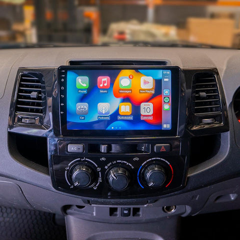 Dashboard of the Toyota Hilux 2005-2015 featuring a multimedia system with Apple CarPlay. The screen displays app icons for Phone, Maps, Messages, Now Playing, Car, Calendar, Settings, and Google Maps. Below the screen are controls for air conditioning and other car functions. The interface indicates a connected iPhone with 4G signal and time display. Type 2